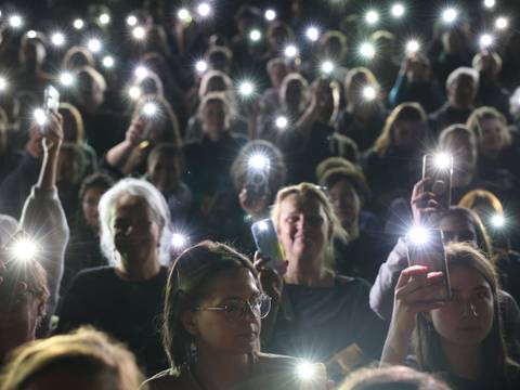  – Ein Blick von der Bühne ins Publikum. Viele Personen im Publikum gucken neugierig und halten ihre Handys mit eingeschalteter Taschenlampe hoch.