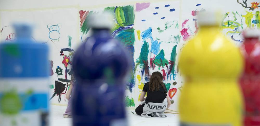 Veranstaltung im Hamburger Bahnhof – Nationalgalerie der Gegenwart – Kind malt vor großem buntem Bild auf Leinwand. Unscharfe bunte Farbflaschen im Vordergrund. Nahe der Leinwand steht NASA auf der Kleidung des Kindes.
