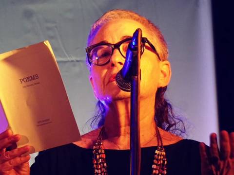 Diti Ronen – Person mit Brille liest in ein Mikrofon und hält ein Buch mit der Aufschrift "POEMS". Im Hintergrund blauer Vorhang.