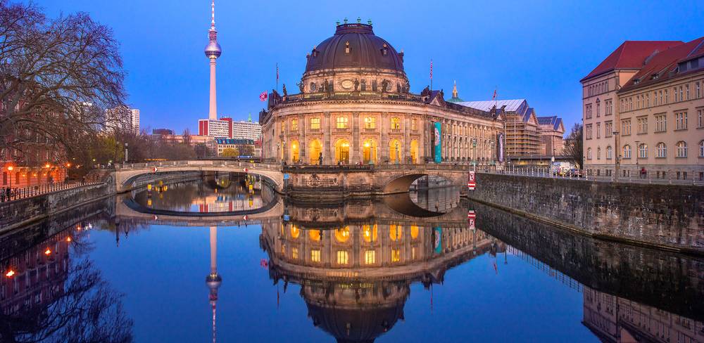 Das Bode-Museum heute – Ansicht der beleuchteten Bode-Museum-Kuppel in Berlin, spiegelnd im Fluss bei Dämmerung. Fernsehturm im Hintergrund, klare, ruhige Wasseroberfläche.