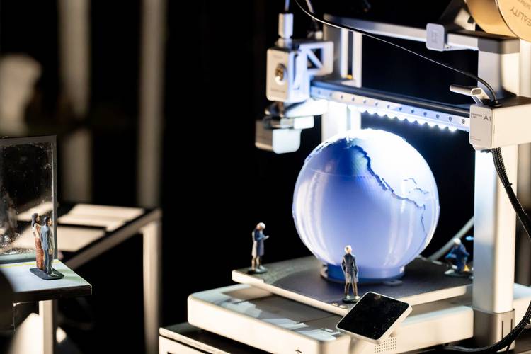 Transport: Fasten your Seatbelts! – 3D-Drucker fertigt blaue Erdkugel an, umgeben von kleinen Figuren, darunter ein Kind. Rechts unten ein Smartphone. Links im Hintergrund ein Spiegel.