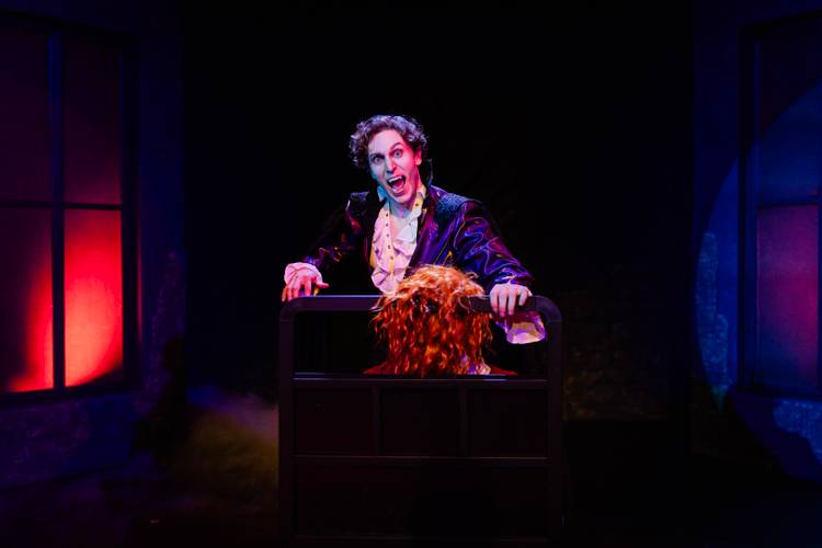 Dracula – A Comedy of Terrors – Person mit blasser Schminke, im weißen Hemd und schwarzem Mantel, steht hinter einer Kiste mit einer roten Perücke. Bühne in blau-rotem Licht.