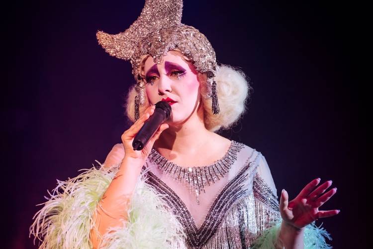 Eine Person mit glitzerndem Kopfschmuck, auffallendem Make-up und Federn am Kostüm singt in ein Mikrofon.