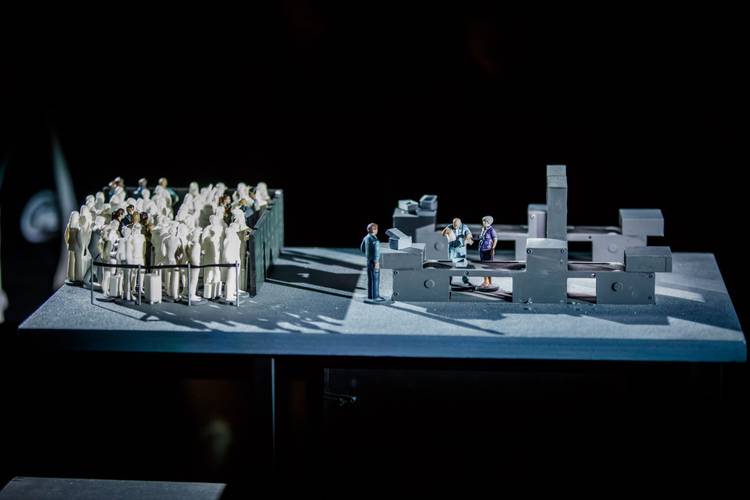 Transport: Connecting Flights – Miniaturfiguren auf geteilter Plattform: links dichte Menschenmenge hinter Geländer, rechts vier Figuren an grauem Bauwerk.