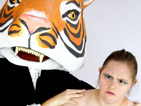  – Person im Tigerkostüm mit großem Kopf hält die Schulter einer Frau. Die Frau trägt ein schulterfreies Kleid in Türkis mit schwarzem Muster.