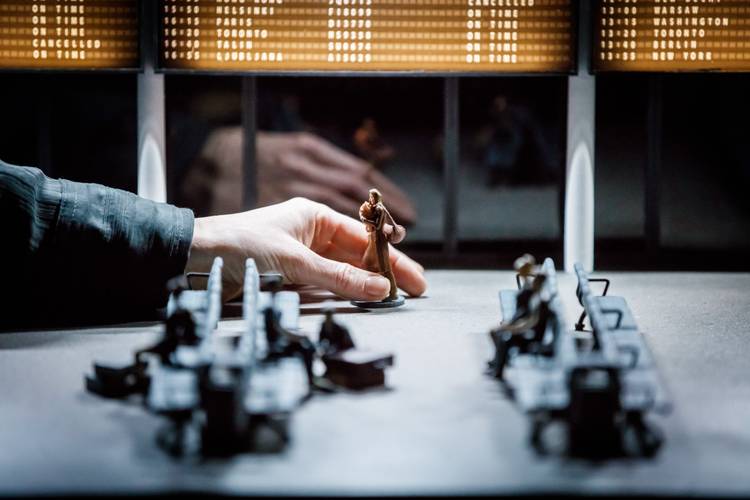 Transport: Connecting Flights – Hand bewegt Spielfigur vor Miniaturbänken, reflektiert in Scheibe. Anzeigetafel im Hintergrund: Lissabon, Warschau, Washington, Toronto, New York.
