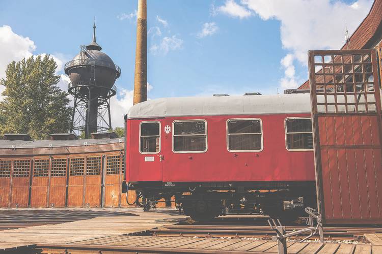Über eine Drehscheibe im Boden und weite Tore lassen sich die Schienenfahrzeuge rangieren und in den Lokschuppen fahren. – Ein roter Abteilwagen steht in der offenen Tür des Lokschuppens. Im Hintergrund erkennt man die Rundung des Schuppens, einen Schornstein und einen Wasserturm.
