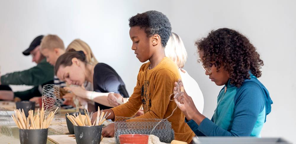Gruppe von Personen in einem Workshop arbeitet an Drahtprojekten an einem Tisch. Erwachsene Personen konzentrieren sich. Im Vordergrund ein Kind in gelbem