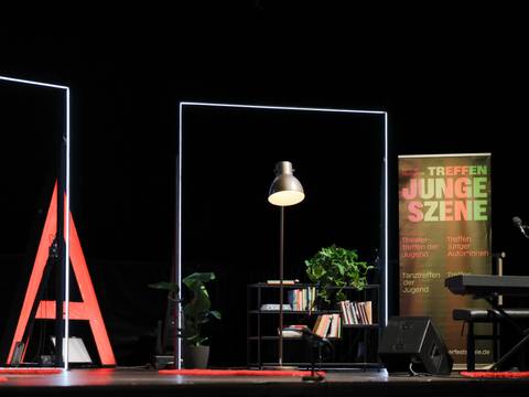Treffen junger Autor*innen – Bühne mit Stehlampe, Pflanzen, Bücherregal. Plakat: Berliner Festspiele, TREFFEN JUNGE SZENE, Treffen junger Autor*innen, Tanztreffen der Jugend.