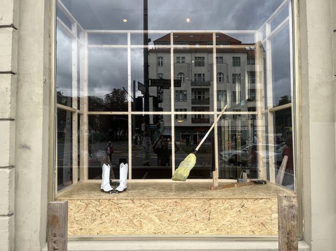 Künstlerinnengespräch »A house and two windows« – Holzkonstruktion in Schaufenster mit Besen, weissen Gummistiefeln und Bauwerkzeug, Spieglung der Strasse und Personen im Hintergrund.