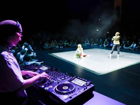 Tanzbegegnungen bei der ARENA – Ein DJ legt an einem Mischpult auf. Im Hintergrund ist ein hell beleuchteter Cypher zu sehen, in dem sich zwei Personen befinden. Das Publikum sitzt rundherum.