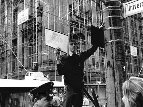 Die Polizei entfernt am 2. Oktober 1994 die vom Aktiven Museum angebrachte Zusatztafel zur Clara-Zetkin-Straße. – Schwarz-Weiß-Fotografie: Ein Polizist entfernt ein zusätzlich angebrachtes Straßenschild des aktiven Museums, während ein weiterer Beamter im Gespräch mit einer Frau steht.