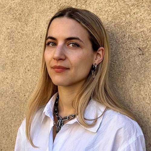 Giulia Giammona – Person mit langem blondem Haar, trägt weiße Bluse und große silberne Halskette, steht vor einer beigen Wand.
