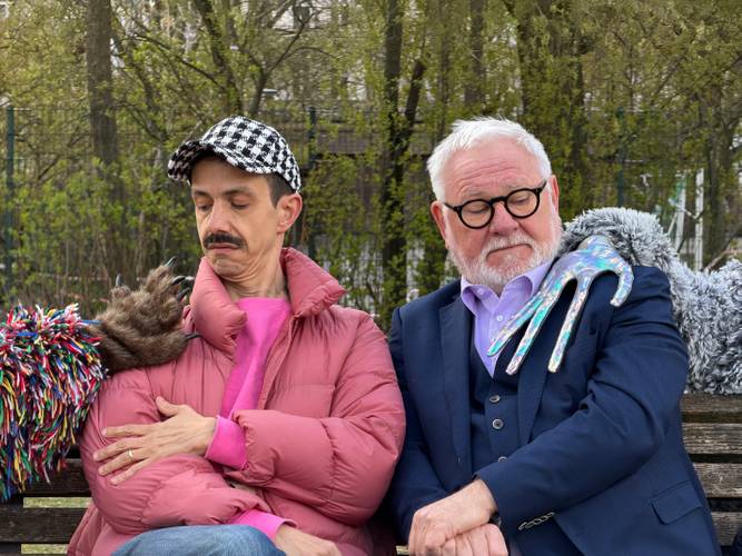 Zwei Personen sitzen auf einer Bank. Person links mit rosa Daunenjacke, bunter Handpuppe und kariertem Hut. Person rechts trägt Anzug, hat grauen Bart.