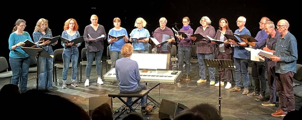 NIE WIEDER! – Person spielt Klavier, umgeben von 14 singenden Erwachsenen in blauer und lila Kleidung, auf einer Bühne stehend, mit Klaviernoten in den Händen.