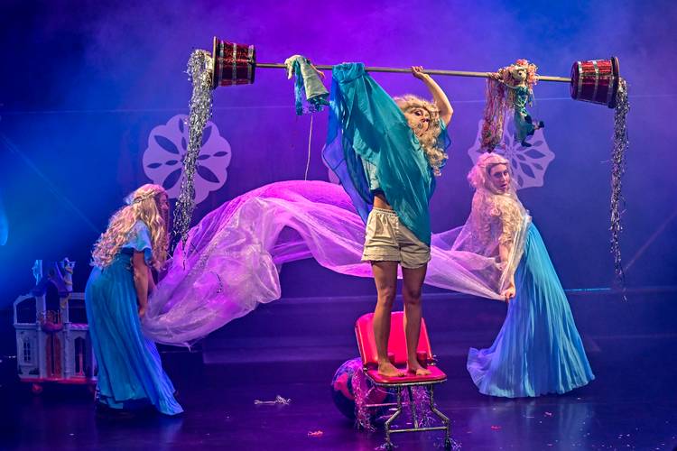 Barbaren Barbies: A wild women circus – Person in blauer Kleidung hebt eine Langhantel mit roten Gewichten auf einer pinken Leiter. Zwei weitere Personen in blauen Kleidern im Hintergrund.