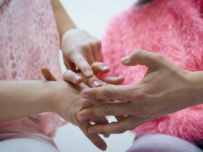 POINT OF TOUCH – Zwei Haende in Interaktion, eine legt sich sanft auf die andere. Personen tragen rosa und pinke Oberteile aus weichem Stoff.