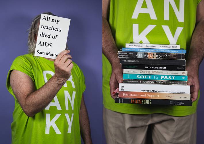 Cranky Choreographing Books – Person mit grauen Haaren in einem grünen T-Shirt mit weißem Schriftzug hält Buch mit Titel "All my teachers died of AIDS Sam Moore" und einen Stapel Bücher.