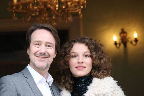 Rainer Bielfeldt und Antonia Bill – Rainer Bielfeldt und Antonia Bill stehen eng nebeneinander und lächeln in die Kamera. Rainer Bielfeldt links trägt ein hellgraues Sakko und ein weißes Hemd, Antonia Bill rechts hat schulterlange lockige Haare, einen dunklen Rollkragen und einen hellen Fellmantel. Im Hintergrund sind ein Kronleuchter und Wandleuchten in einem Innenraum zu sehen.