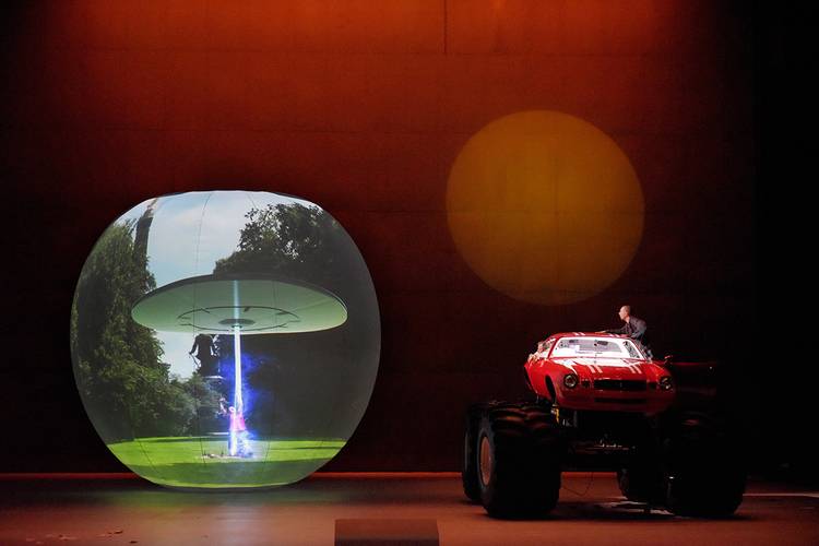 Die Entführung aus dem Serail – Großes, beleuchtetes Objekt mit UFO-Bild; rechts rotes Auto mit großen Reifen, Person auf dem Dach; Hintergrund in Orange gehalten.