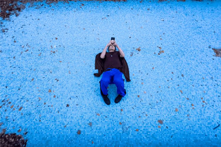 Person liegt auf dem Rücken auf blauem Boden, trägt blaue Hose, schwarzen Pulli, schwarze Schuhe und hält ein Smartphone nach oben gerichtet.