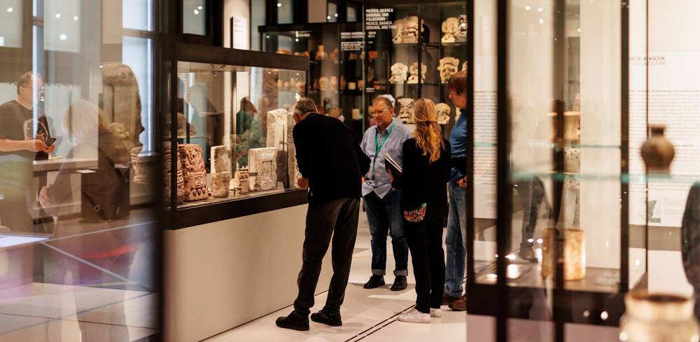 Besucher*innen einer Führung im Bereich "Grafische Kommunikationssysteme in Mesoamerika" des Ethnologischen Museums – Personengruppe betrachtet antike Artefakte in einer modernen Museumsvitrine. Im Hintergrund weitere Ausstellungsstücke hinter Glas.