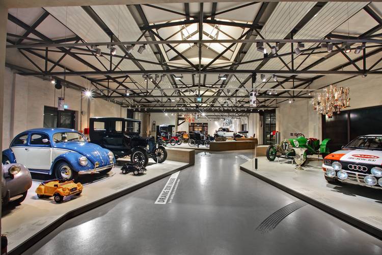 Elf Themeninseln geben Einblicke in die Welt der Mobilität. – Blick in die Straßenverkehr-Ausstellung: Eine große Halle mit Stahlkonstruktion an der Decke. Ein breiter Gang zwischen niedrigen Podesten mit verschiedenen Fahrzeugen und Objekten.
