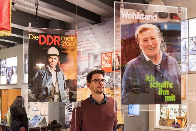 Dauerausstellung "Alltag in der DDR" – ein Mann mit rotem Pullover und schwarzer Brille betrachtet Propagandaplakate, die in der Ausstellung "Alltag in der DDR" ausgestellt werden