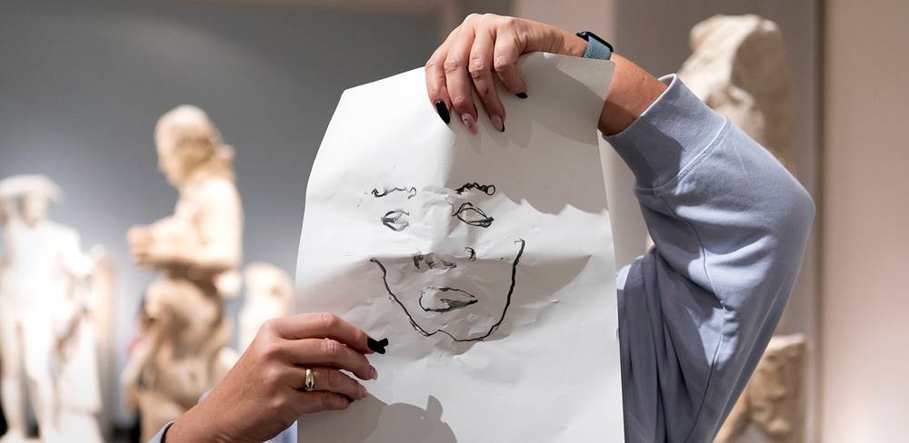 Workshop im Alten Museum – Person hält Blatt Papier mit Gesichtszeichnung; verschwommene Skulpturen im Hintergrund; Fotografie in Innenraum.