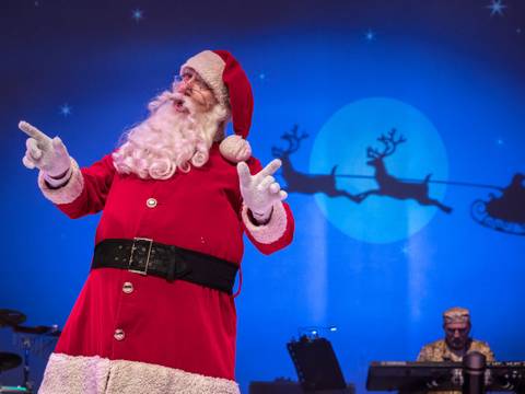Das Märchen von der Prinzessin und dem Weihnachtsmann – Person in Weihnachtsmannkostüm vor einer blauen Kulisse mit Rentierschlitten-Silhouette. Im Hintergrund eine Person an einem Keyboard.