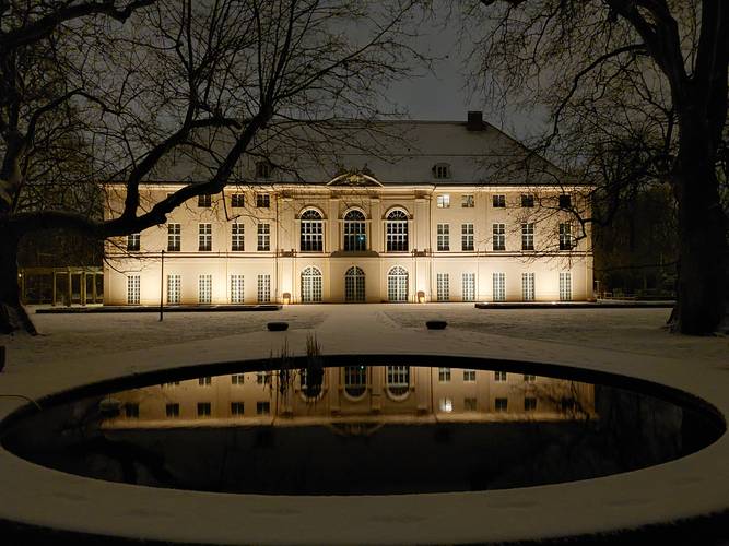 Schloss Schönhausen im Winter – Ein großes, nachts beleuchtetes Gebäude mit symmetrischer Fassade und hohen Bogenfenstern, umgeben von kahlen Bäumen, deren Spiegelbild sich in einem runden Teich im Vordergrund widerspiegelt: Schloss Schönhausen im Winter