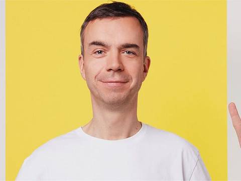  – Person in weißem T-Shirt vor gelbem Hintergrund zeigt mit rechter Hand das Peace-Zeichen.