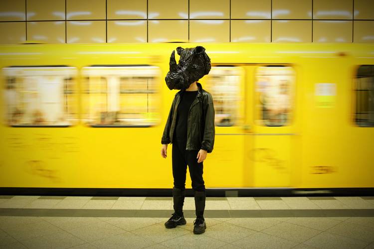 Rhinozeros – ein Comeback – Person mit schwarzem Helm in Form eines Dino-Kopfes steht vor gelber U-Bahn im U-Bahnhof. Zug in Bewegung. Person in dunkler Kleidung und Stiefeln.