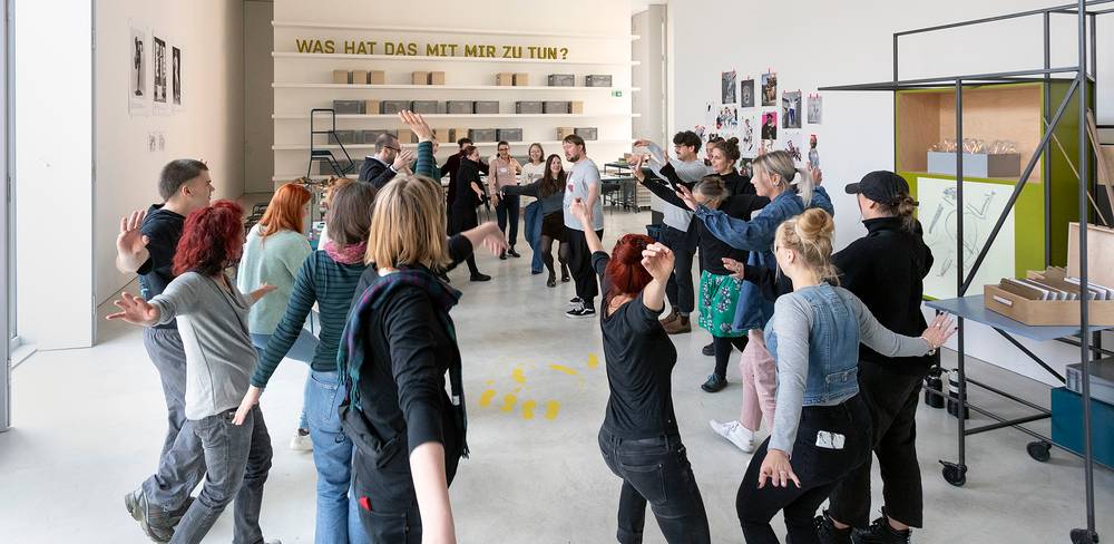 Menschen in Kreisformation, erhobene Arme. An der Wand der Schriftzug: "WAS HAT DAS MIT MIR ZU TUN?" Raum mit Kunstwerken und Regalen im Hintergrund.