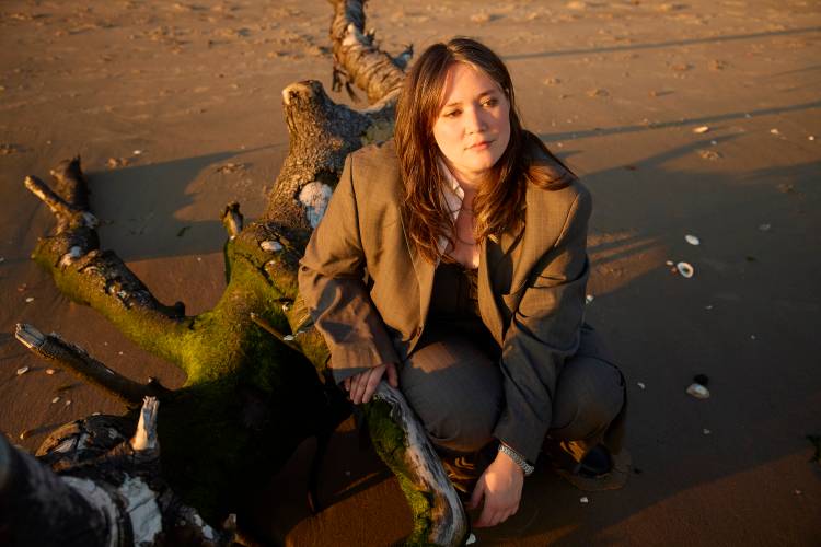 Wendy Eisenberg – Weiblich gelesene Person mit braunen, offenen Haaren und grauem Blazer vor einer Strandlandschaft bei Sonnenuntergang.