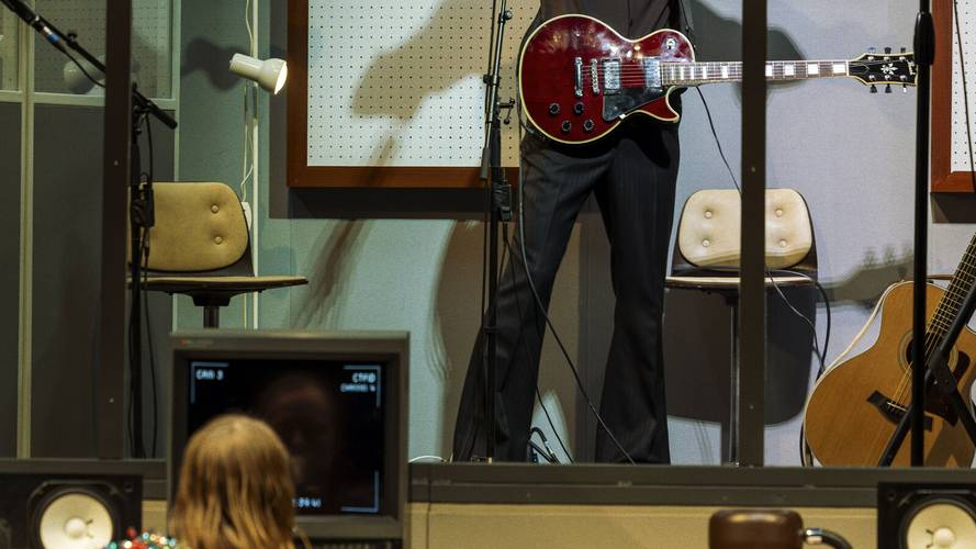 Stereophonic – Person steht mit roter E-Gitarre hinter Glaswand; Person vorm Fernseher mit Reflexion. Zwei beige Stühle, eine braune Akustikgitarre im Raum.