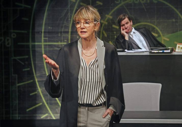 Person mit Brille und gestreifter Bluse spricht gestikulierend; zweite Person sitzt im Hintergrund an einem Tisch, gestützt den Kopf mit der Hand.