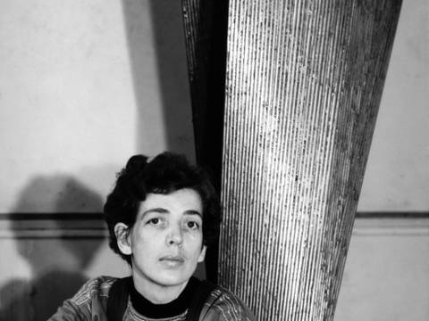 Brigitte Meier-Denninghoff – Schwarz-weiß-Foto: Brigitte Meier-Denninghoff sitzt neben einer, zum Teil abgebildeten, silbernen Skulptur am Boden. Die Arme leicht übereinander auf die Knie gestützt, blickt sie direkt in die Kamera.