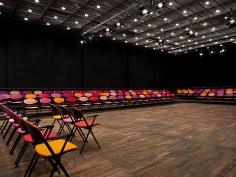  – Leerer Veranstaltungssaal mit vielen Stuhlreihen in Rot-, Violett- und Orangetönen auf einem Holzboden, Decke mit Lampen.