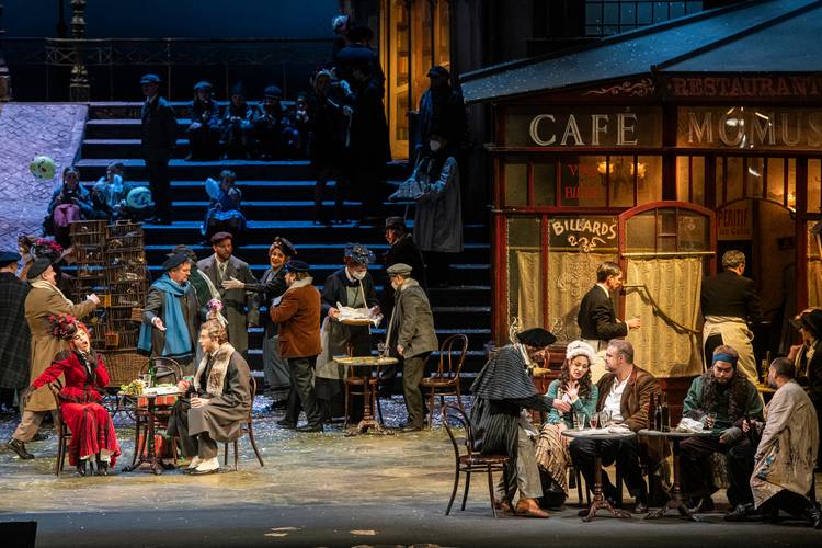 La bohème – Opernszene mit Café Momus; viele Personen sitzen oder stehen, einige mit Getränken. Bühne im Freien mit winterlicher Atmosphäre.