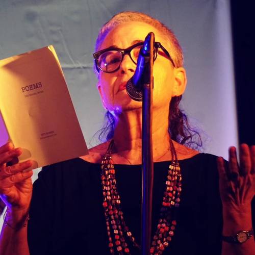 Diti Ronen – Person mit Brille liest in ein Mikrofon und hält ein Buch mit der Aufschrift "POEMS". Im Hintergrund blauer Vorhang.