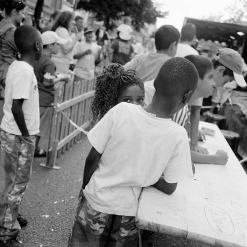 Akinbode Akinbiyi, Wedding, Berlin, 2005, Aus der Serie: „African Quarter“, seit den 1990er Jahren – Schwarz-weiß-Fotografie: Im quadratischen Format zeigt die Fotografie mehrere Personen, die vorwiegend mit dem Rücken zur Kamera stehen, während eine junge, weiblich gelesene Person direkt in die Kamera blickt.