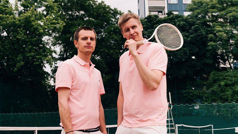 Zwei Personen in rosa Polohemden auf einem Tennisplatz; eine hält einen Tennisball, die andere einen Schläger auf der Schulter; Bäume und Gebäude im