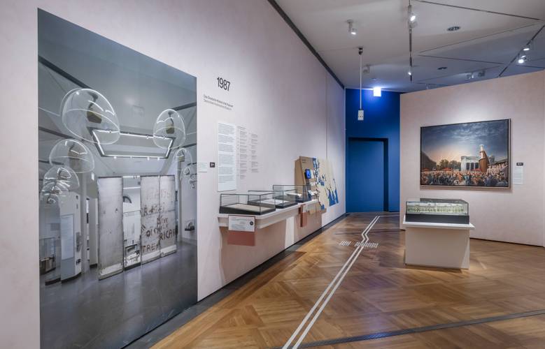Raumansicht der Ausstellung „Objekte. Geschichte. Geschichten. Blick in die Sammlung“. Auf der linken Seite ist ein etwa zwei Meter hohes Foto einer früheren Ausstellung des DHM zu sehen. Dahinter sind drei kleine Vitrinen sowie eine interaktive Landkarte zu erkennen. Rechts im Bild ist ebenfalls eine Vitrine im Raum zu erkennen, dahinter hängt ein Gemälde.
