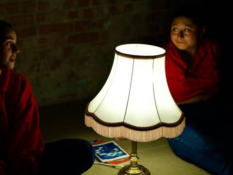  – Zwei Personen in roten Hemden sitzen im Dunkeln neben einer eingeschalteten Tischlampe und einem Notizbuch auf dem Boden.