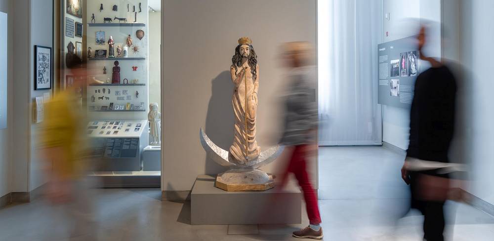 Ansicht der Dauerausstellung „Kulturkontakte. Leben in Europa“ – Skulptur einer stehenden Figur mit Krone, umgeben von einem Halbmond. Verschwommene Menschen bewegen sich seitlich; Hintergrund: Museumsvitrine.