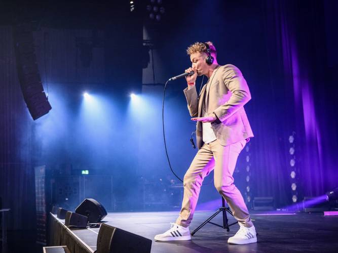 BEATHOVEN – Beat für Elise – Eine Person in beigefarbenem Anzug und weißen Sneakers singt mit Mikrofon auf der Bühne, umgeben von blauem und violettem Licht, trägt Kopfhörer.