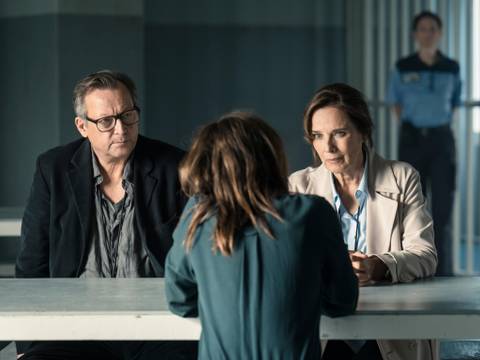 Matthias Brand und Désirée Nosbusch. © Stephan Rabold – Drei Leute sitzen an einem Tisch, hinter ihnen ein Polizeibeamter.