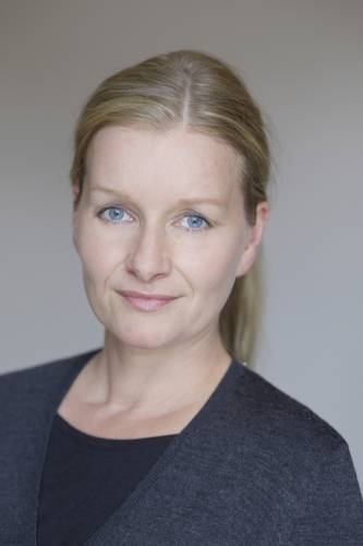 Julia Encke – Person mit blonden Haaren und blauer Strickjacke blickt direkt in die Kamera, neutraler Gesichtsausdruck, grauer Hintergrund.