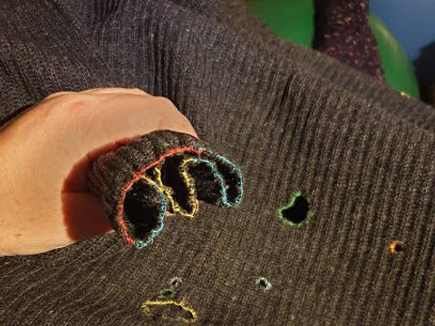  – Eine Hand im grauen Strickpullover mit bunten Saumkanten. Mehrere sichtbare Loecher im Pullover. Im Hintergrund ein gruener und ein blauer Gegenstand.
